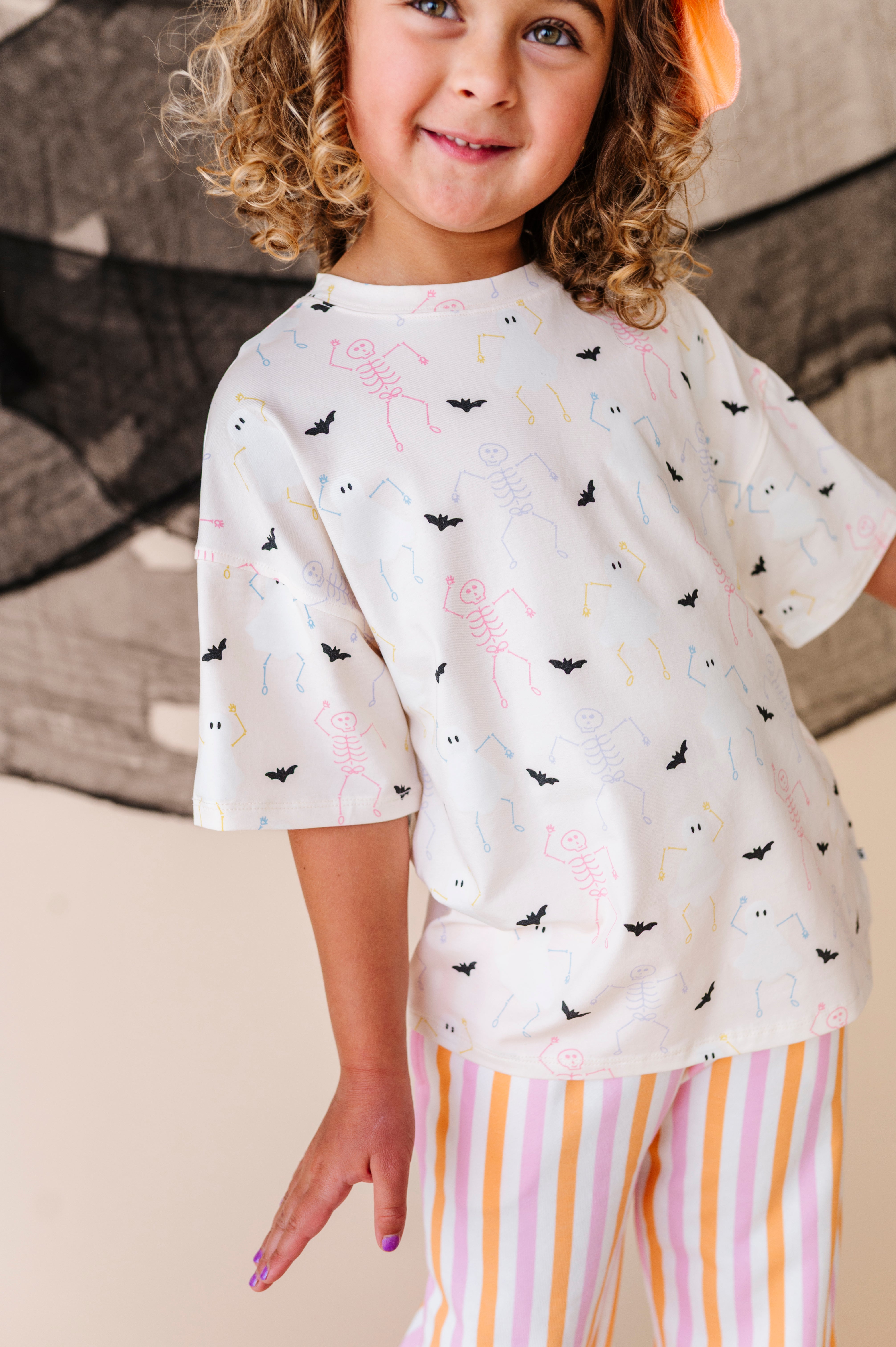 Child wearing a cream halloween themed t-shirt with colorful patterns and striped pants, sitting on a couch.